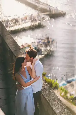 Man kiss on the forehead his girlfriend while they hug each other leaning over a wall with Sorrento beach in the background
