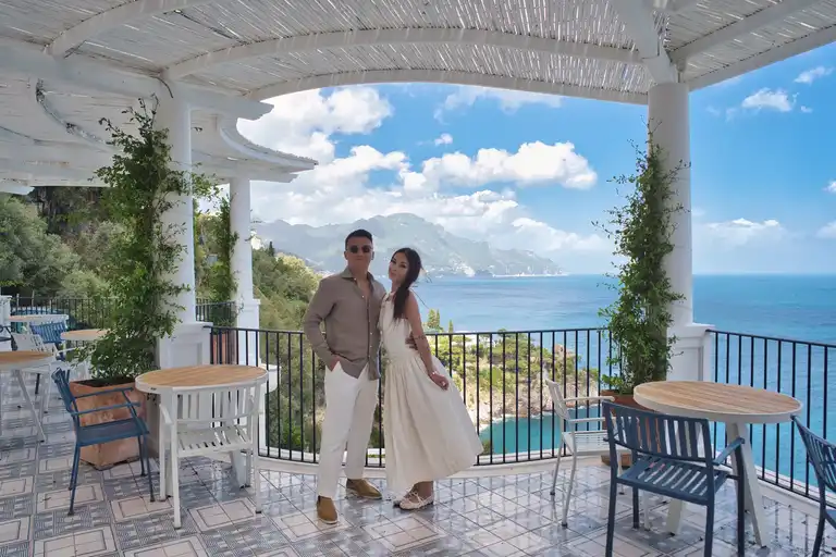 A candid, joyful close-up of the newlyweds laughing together while walking hand-in-hand along a stone pathway that wraps around the rugged cliffside of Borgo Sant'Andrea near Amalfi.