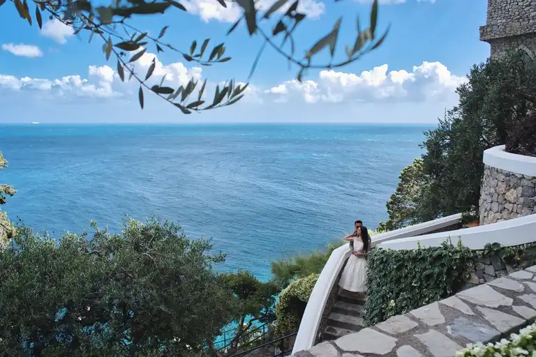 A candid moment of the newlyweds holding each other, with the vertical, rugged cliff face of Borgo Sant'Andrea rising dramatically behind them and the blue sea to their side.