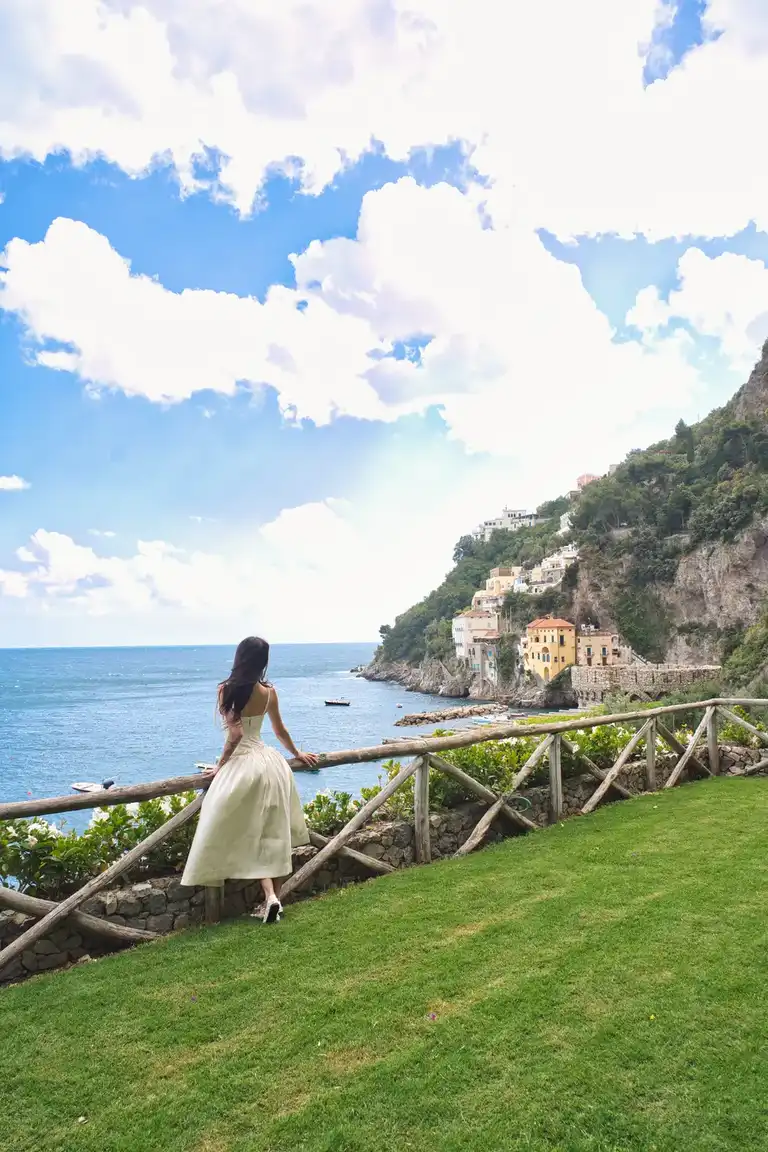 A back view portrait of the bride, in her white wedding dress, standing alone on a lower terrace path of Borgo Sant'Andrea, with the massive cliff face looming over her and the sea visible in the distance.