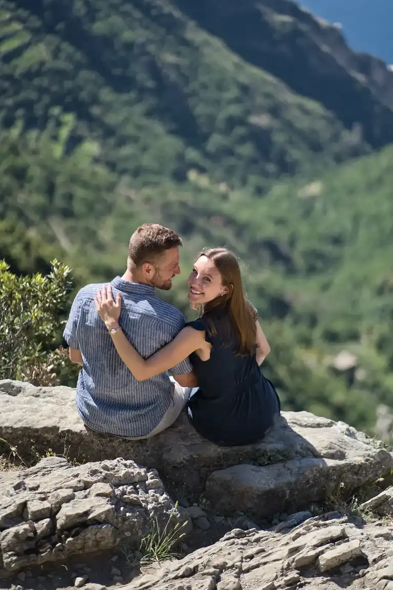Relaxed newly engaged couple sitting on a cliff edge along the Path of the Gods hiking trail, admiring the engagement ring at sunrise near Bomerano, Amalfi Coast.