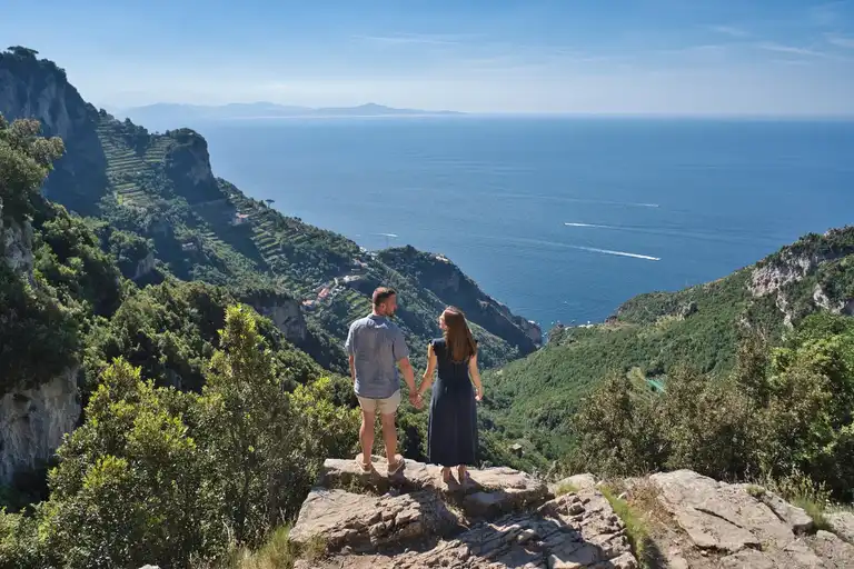 Newly engaged couple standing on a mountain trail overlooking a sweeping panoramic view of the sea and mountains during a sunrise hike on the Sentiero degli Dei (Path of the Gods) from Bomerano.