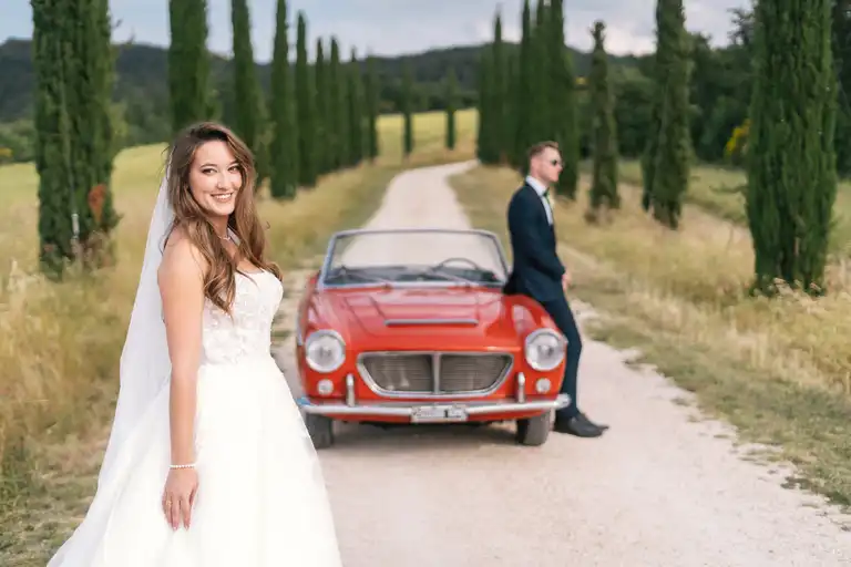 Bride pose in front while broom sits on old vintage italian car with typical tuscany cypresses in the background